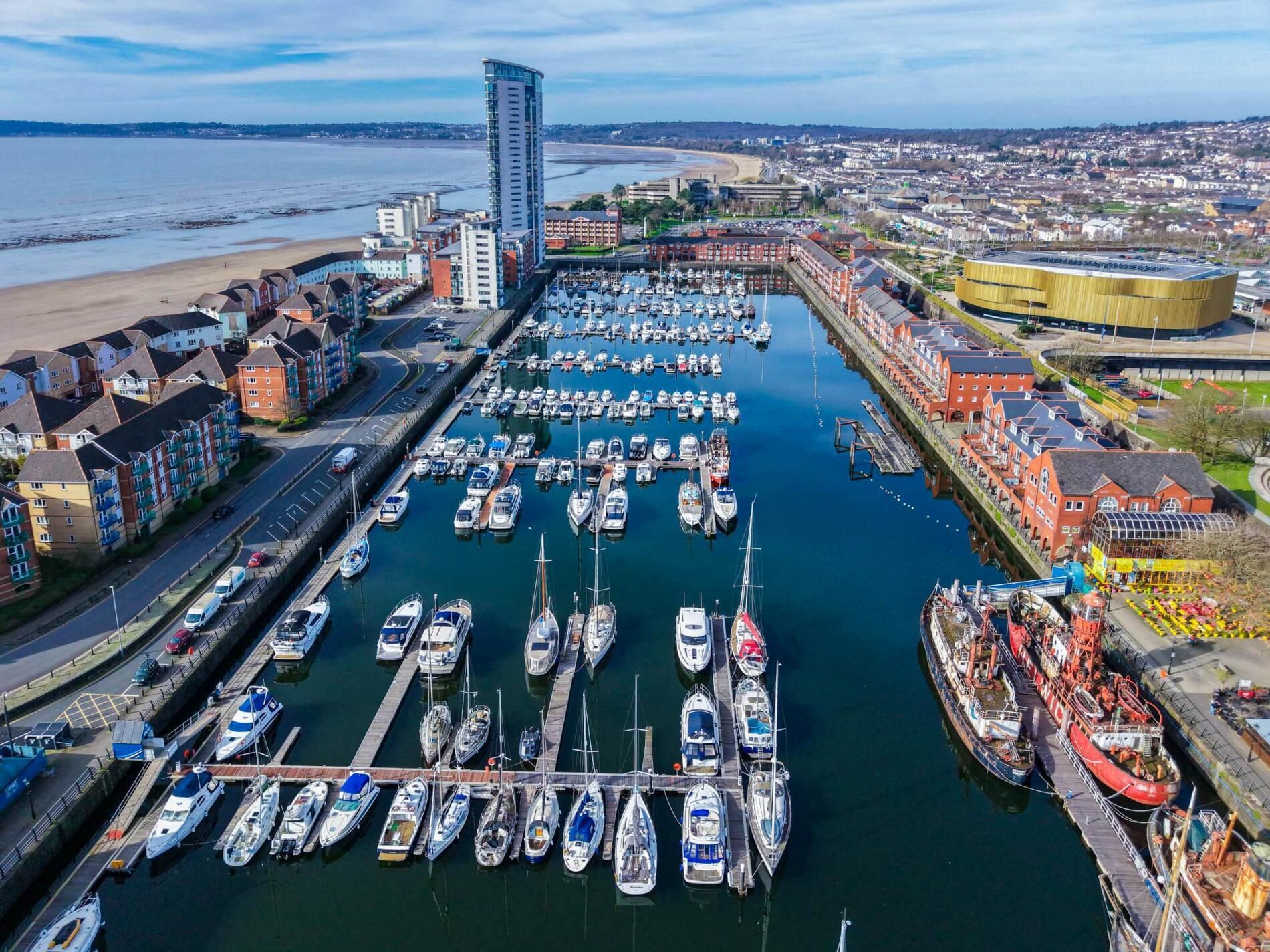 Un port de plaisance où sont amarrés de nombreux bateaux, un grand bâtiment sur la gauche et un stade sur la droite.