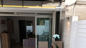 En uteplats med ett bord och stolar, en glasdörr som leder in till ett inomhusutrymme och en liten trädgårdsdel med en växt.