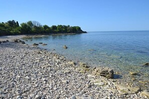 A pebbled beach with clear water, a rocky shoreline, and a distant tree line. - Holiday apartment with terrace (Novigrad)