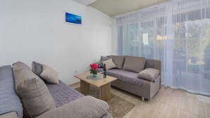 Ett vardagsrum med en soffgrupp, ett litet bord med en vas och ett stort fönster med tunna, genomskinliga gardiner.