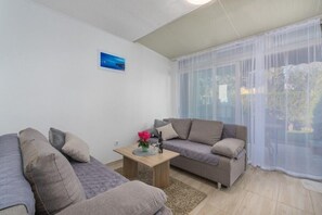 Ein Wohnzimmer mit einer Sofagarnitur, einem kleinen Tisch mit einer Vase und einem großen Fenster mit transparenten Vorhängen.