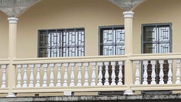 Eine Gebäudefassade mit drei Fenstern, jedes versehen mit Metallgitter, und einem Balkon mit weißen Brüstungen.