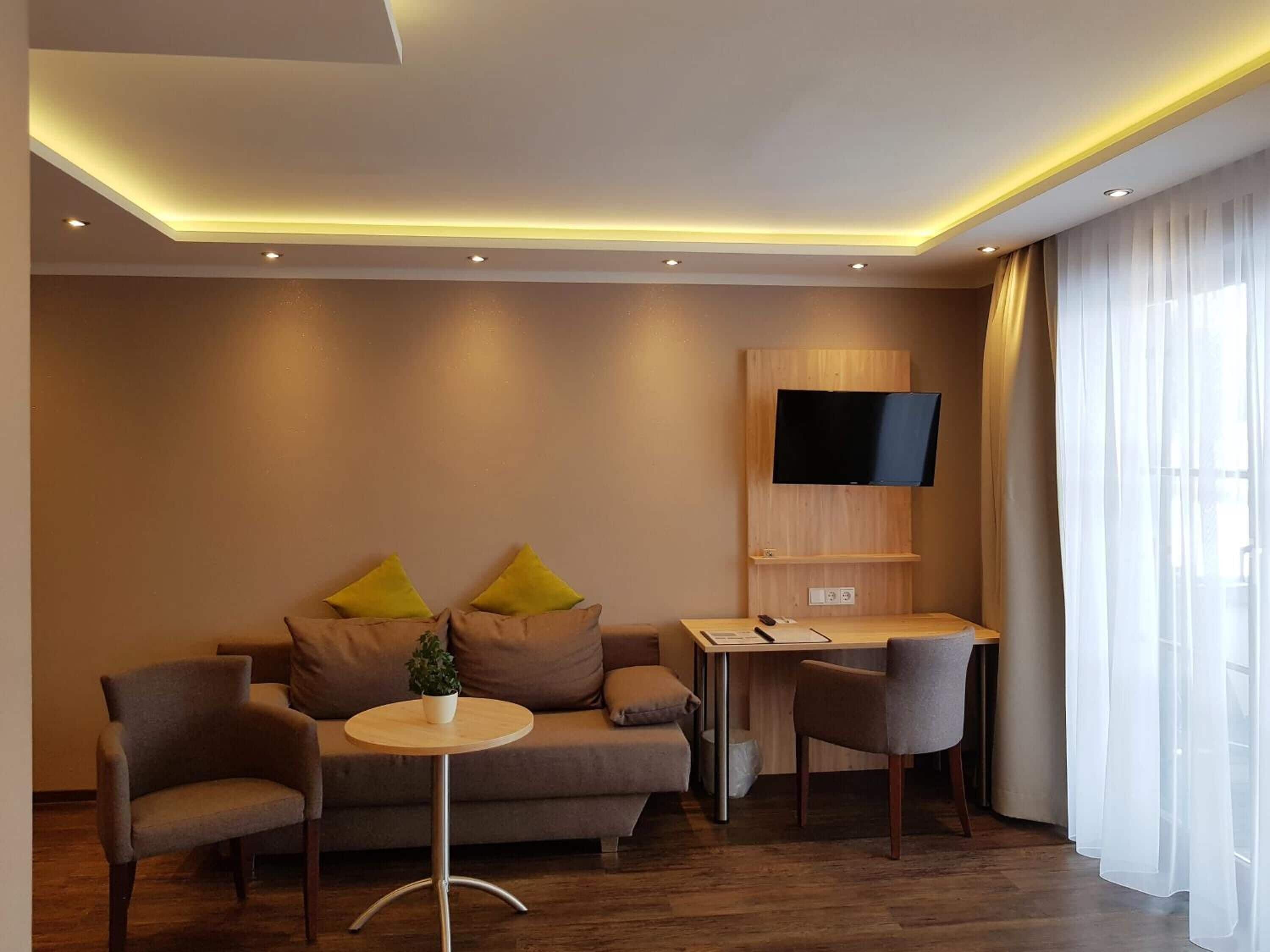 A modern hotel room with a sofa, two armchairs, a small round table, a desk, and a mounted TV.