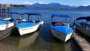 A row of small boats docked at a pier with clear water and mountains in the background. - Apparthotel Garni Superior Simsseeblick (Bad Endorf)