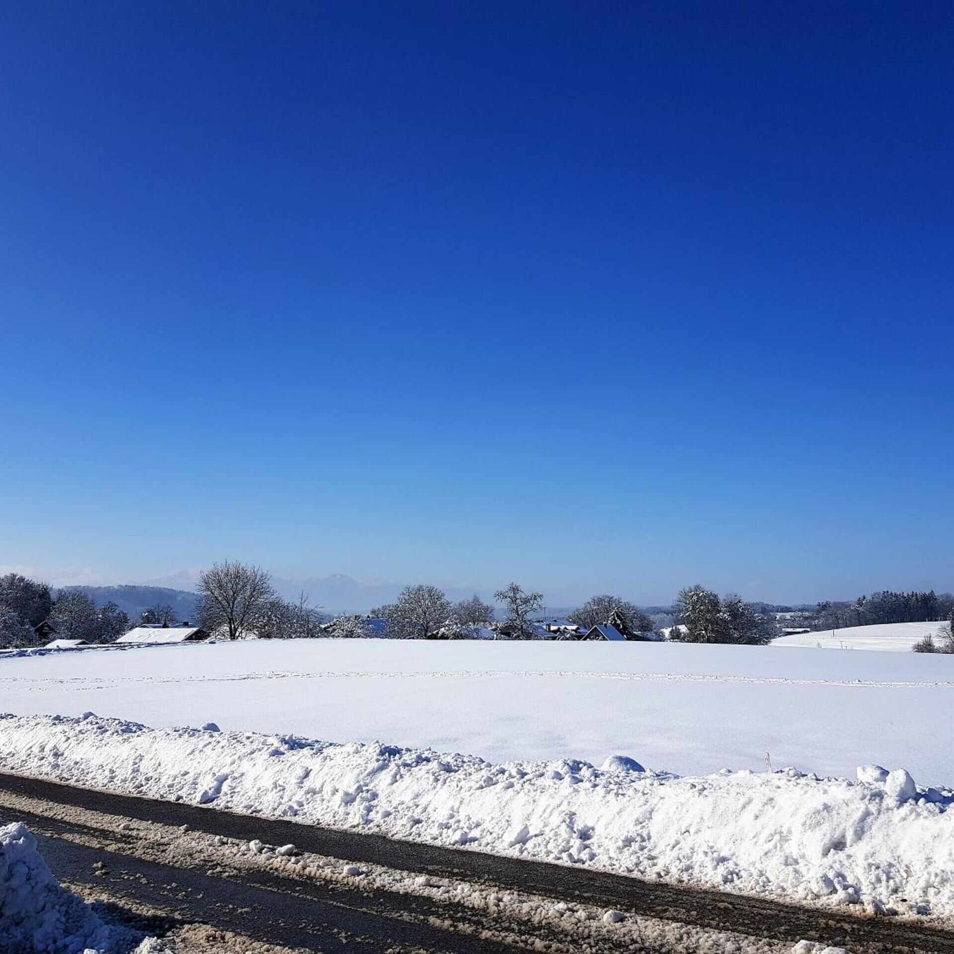 Een besneeerd landschap met een helderblauwe lucht, een weg en gebouwen in de verte.