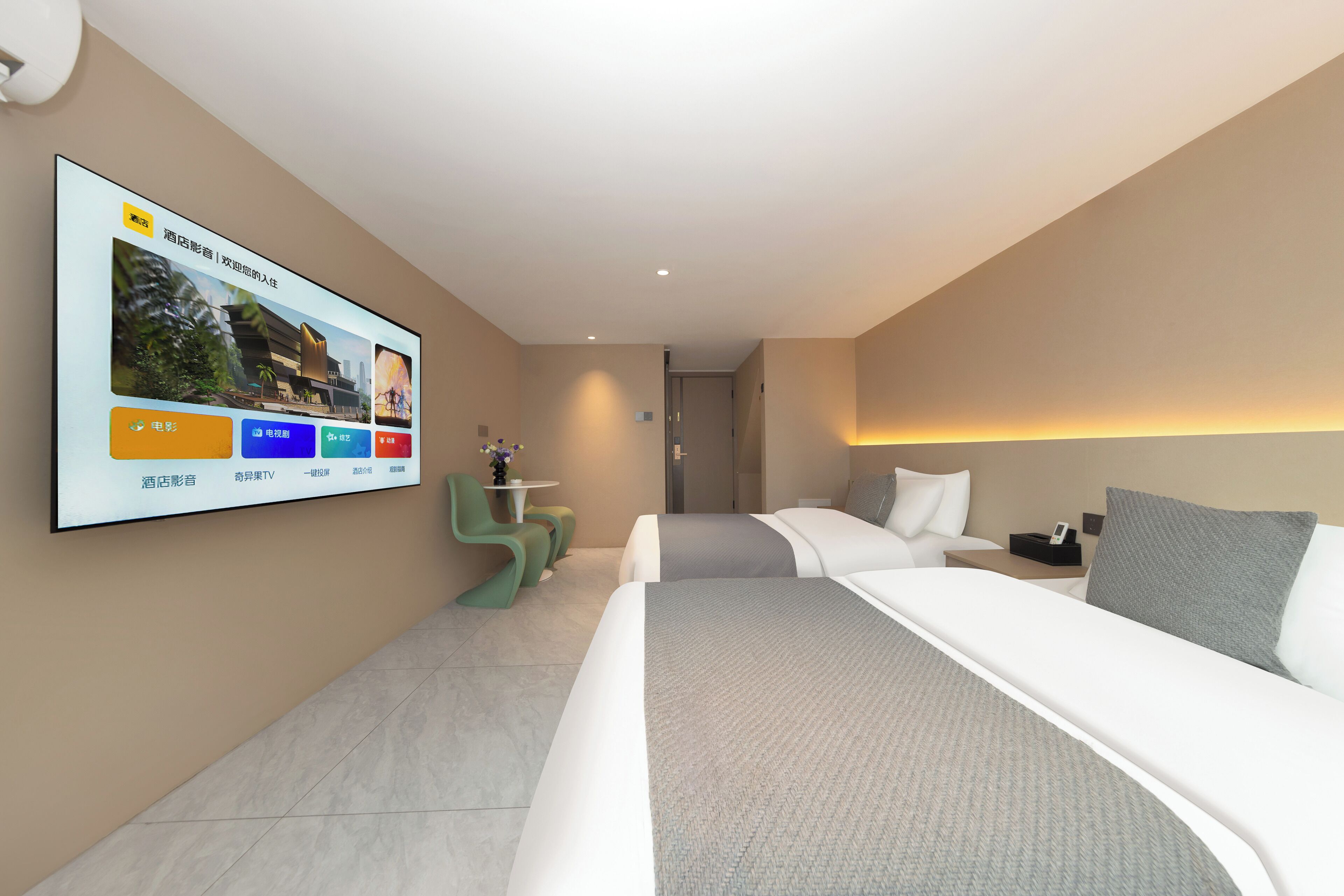 Ett modernt hotellrum med en stor TV som är monterad på väggen, en säng med vita sängkläder, ett litet bord med en vas med blommor och en sittgrupp med gröna stolar.