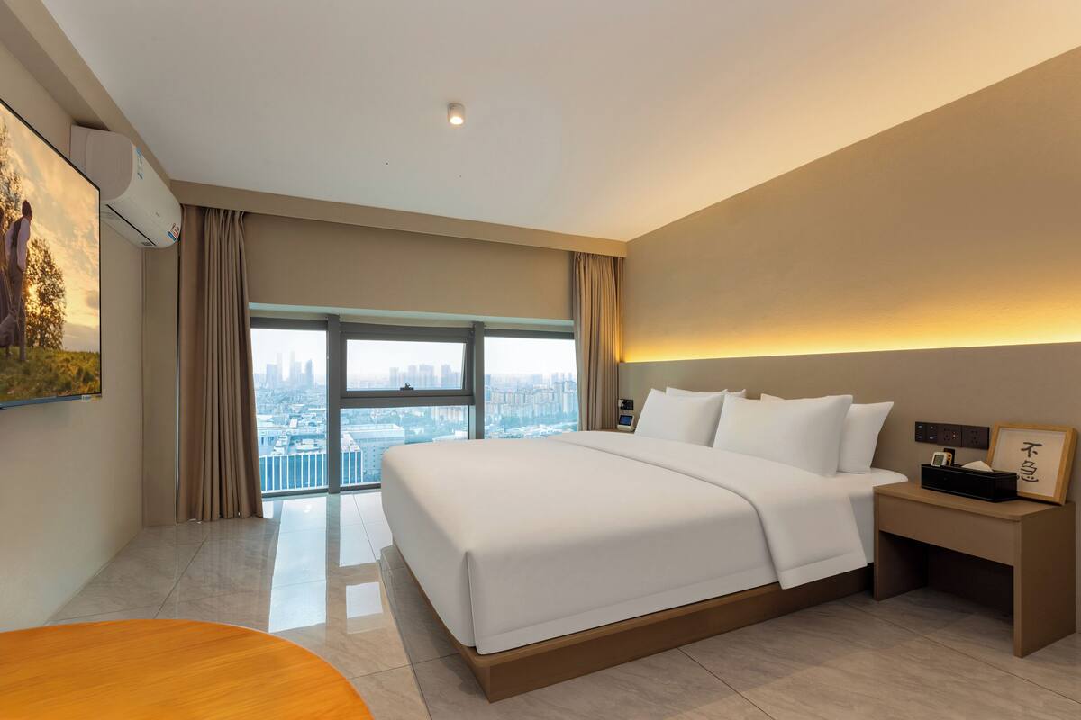 Habitación de hotel moderna con una cama grande, una mesita de noche, aire acondicionado en la pared y vistas a la ciudad a través de amplios ventanales.