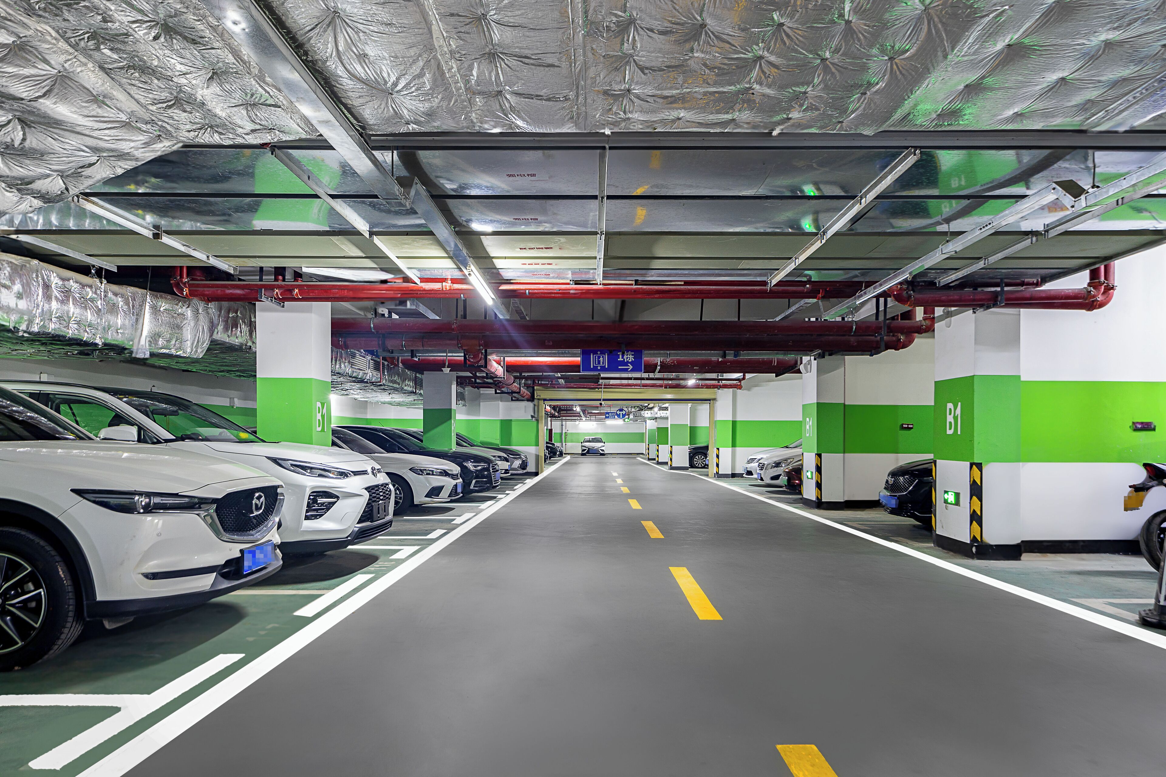 A parking garage with multiple rows of parked cars, green and white walls, and overhead pipes.