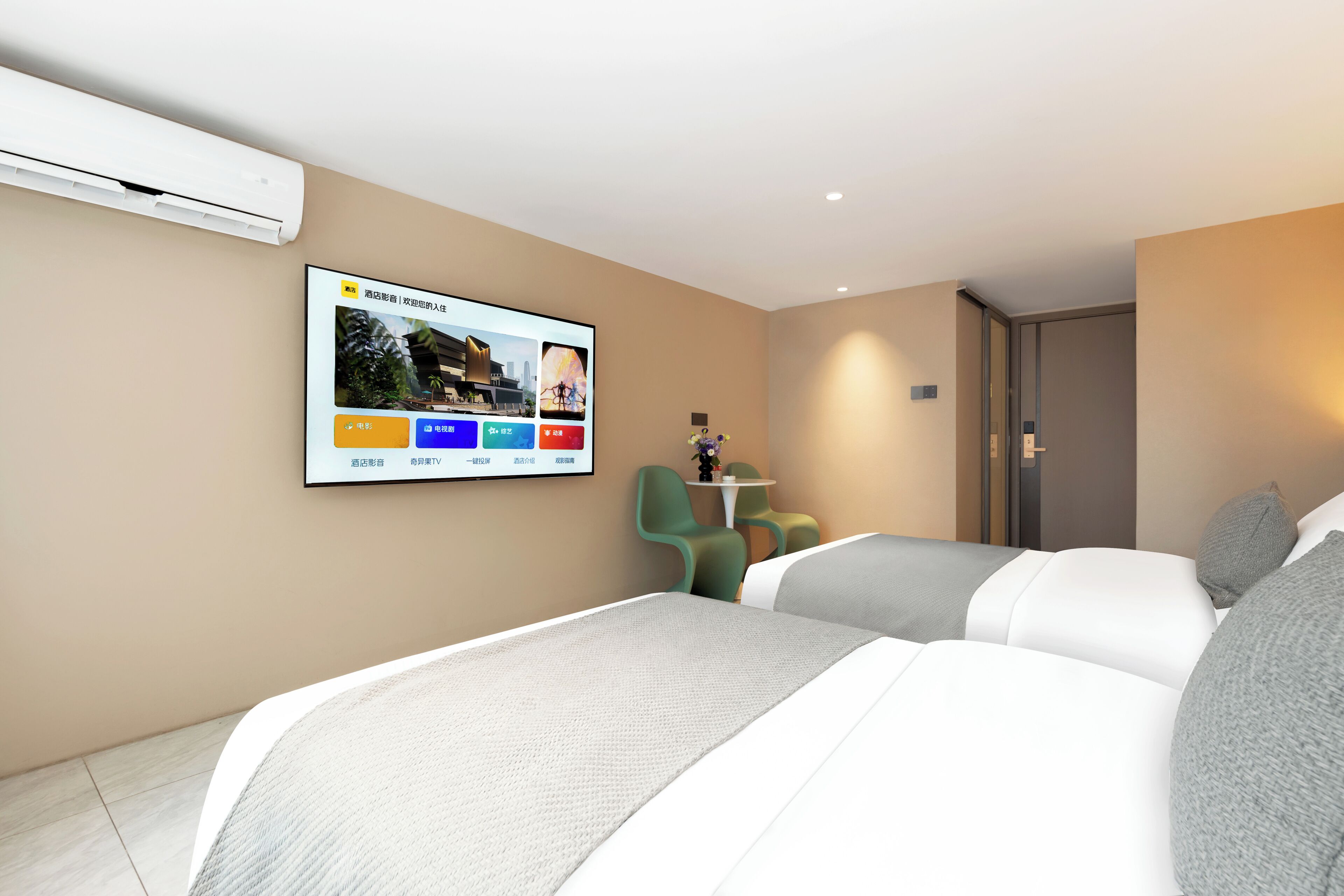 A hotel room with a bed, a TV mounted on the wall, a small table with chairs, and an air conditioning unit.