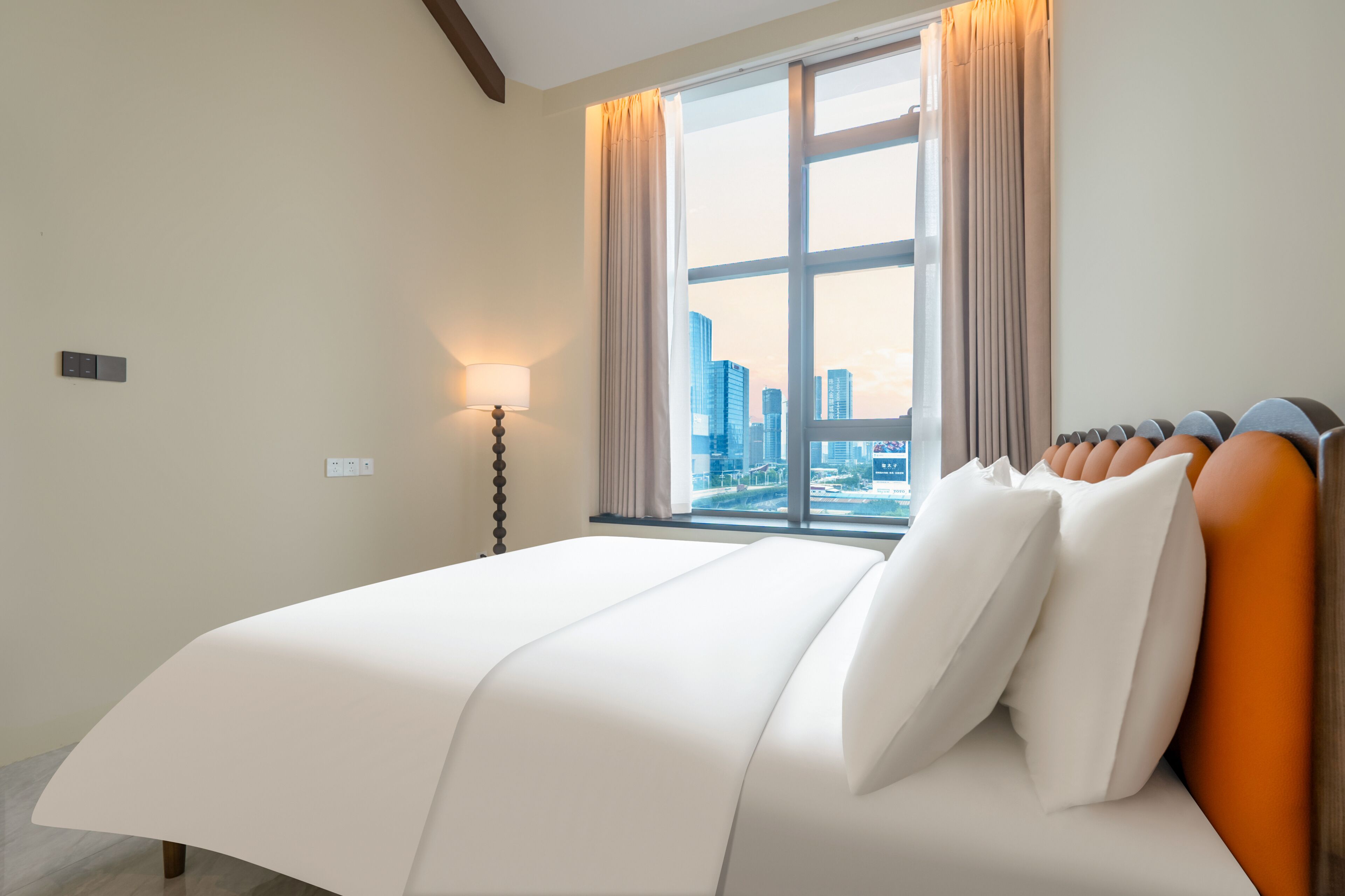 A hotel room with a large bed, a nightstand, a lamp, and a view of a cityscape through a large window.