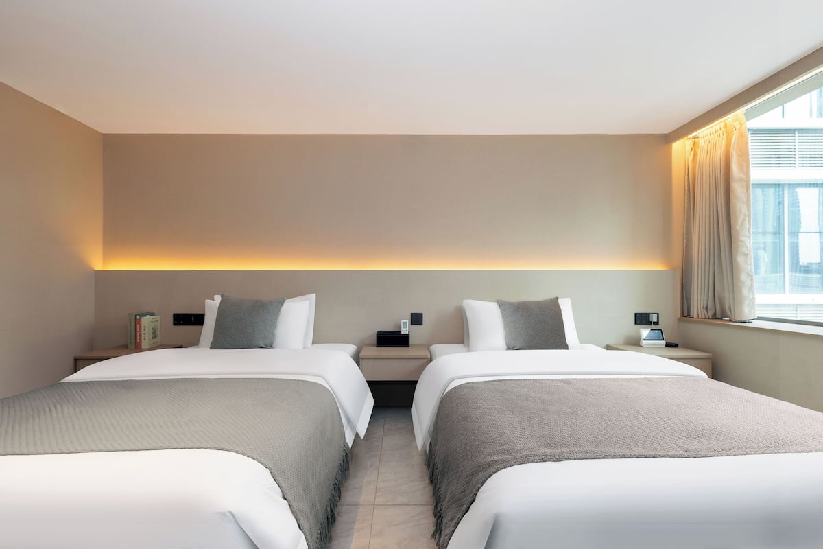 Habitación de hotel con dos camas, una mesita de noche con lámpara, una pequeña mesa con teléfono y una ventana con cortinas.