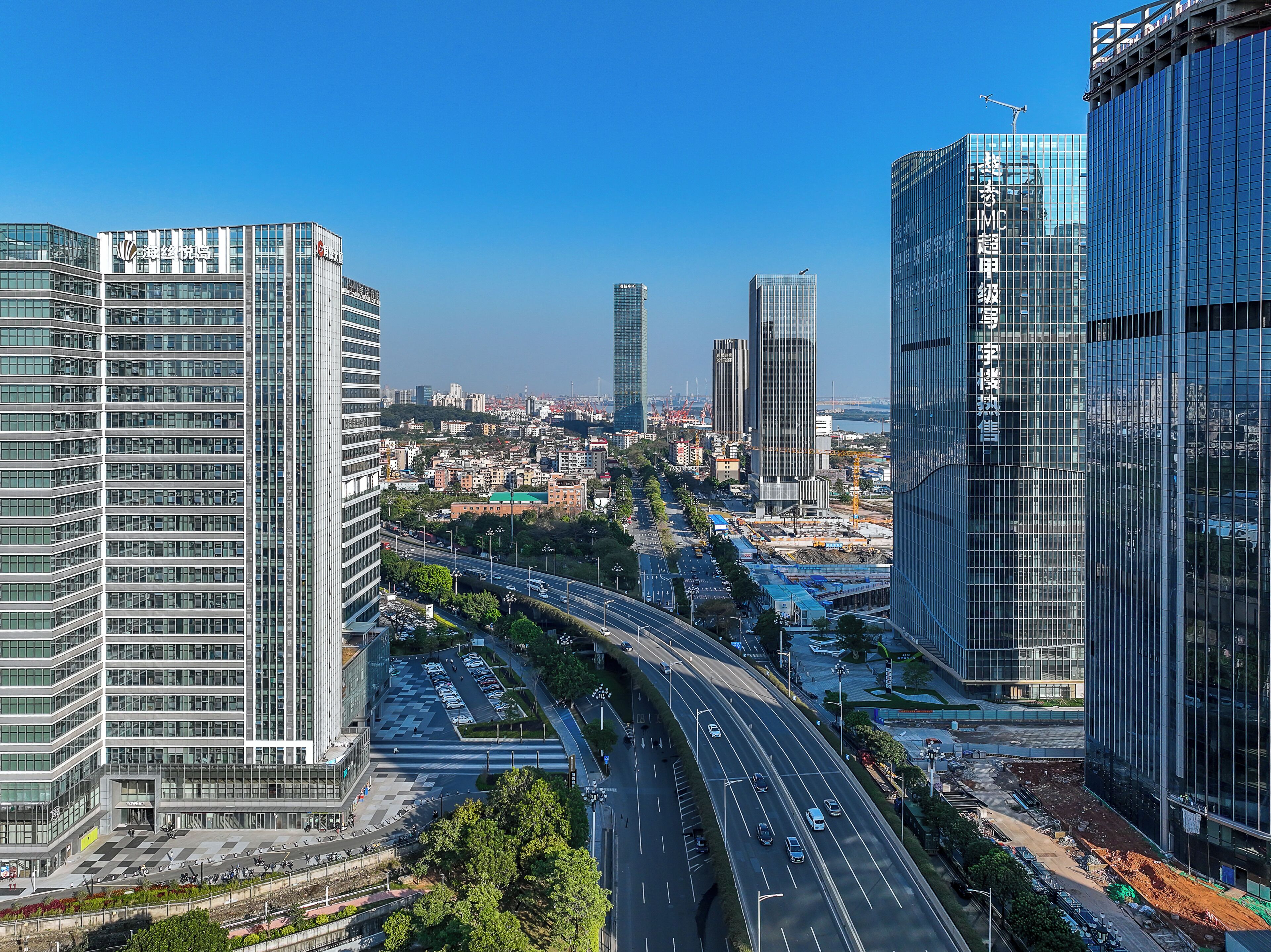 A cityscape with modern high-rise buildings, a multi-lane road, and a clear blue sky.