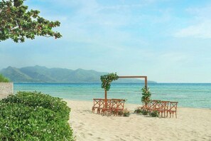 Decoració de boda a la platja, amb arc de fusta i cadires de vimet, envoltat de vegetació i vistes al mar i a les muntanyes.