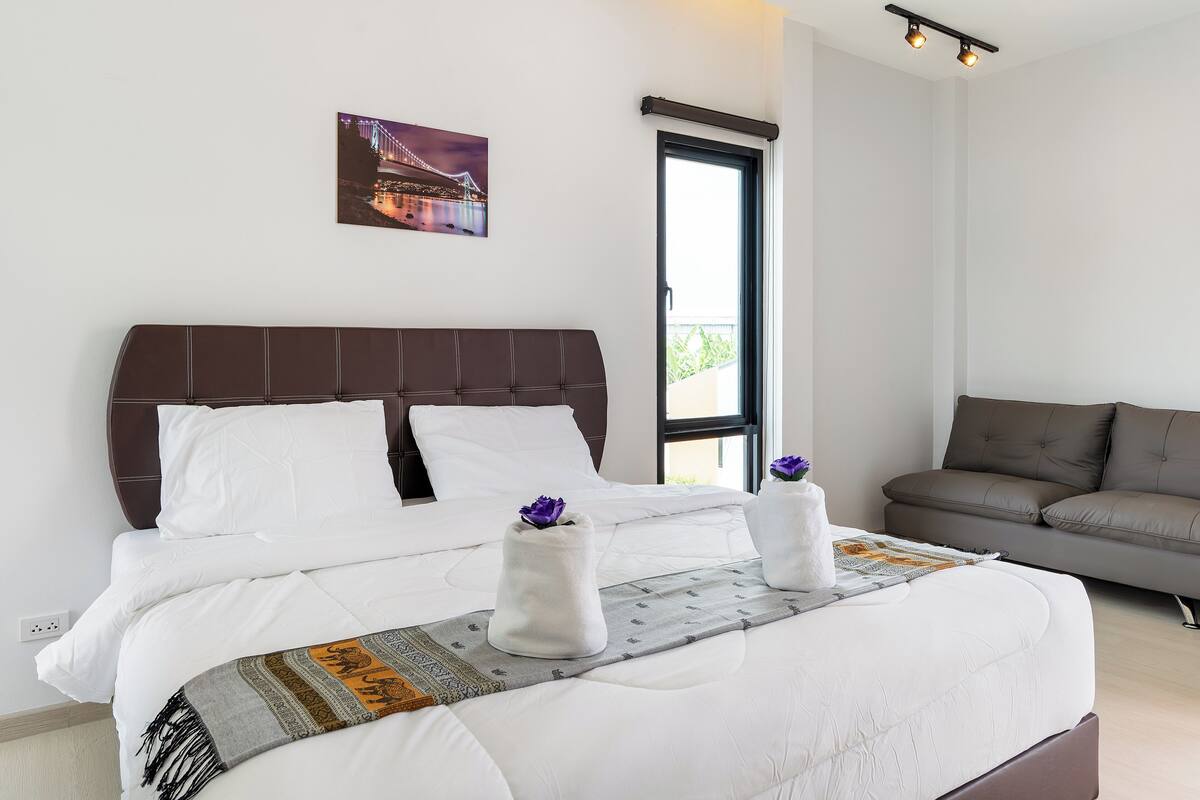 A neatly made bed with white linens, a brown headboard, and two white towels with purple flowers. There is a gray throw blanket with a patterned design, a gray pillow, and a framed picture on the wall.