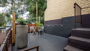 En udendørs patio med støbt gulv, træhegn, en metalstol, en høj plantekasse og en trappe op til en bygning med murstens- og sort facade.