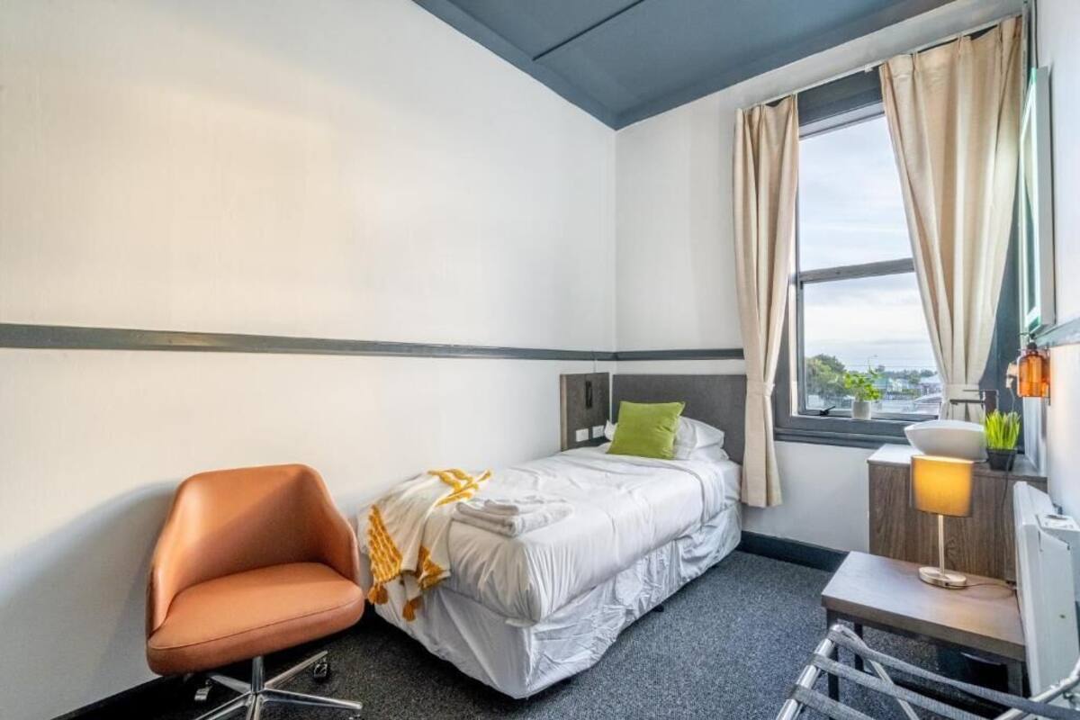A hotel room with a bed, a chair, a desk, a lamp, and a window with curtains.
