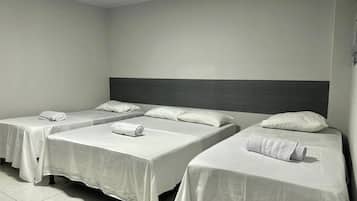 A hotel room with three single beds, each with a white bedspread and a rolled towel. The room has a simple, clean design with a dark headboard and white walls.