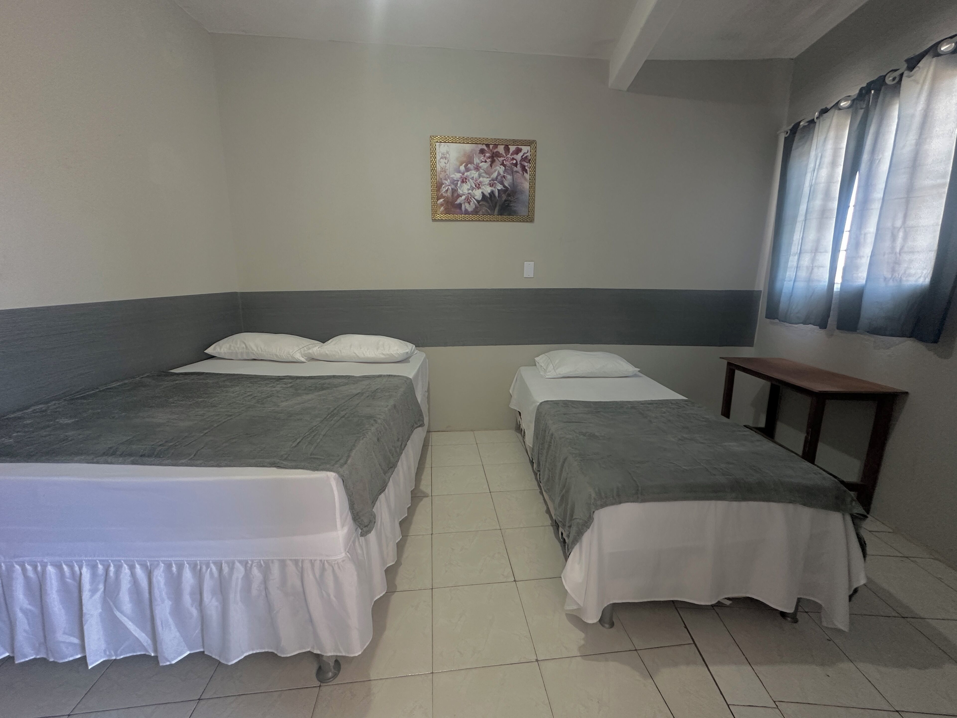 A room with two beds, a small table, a painting on the wall, and a window with curtains.