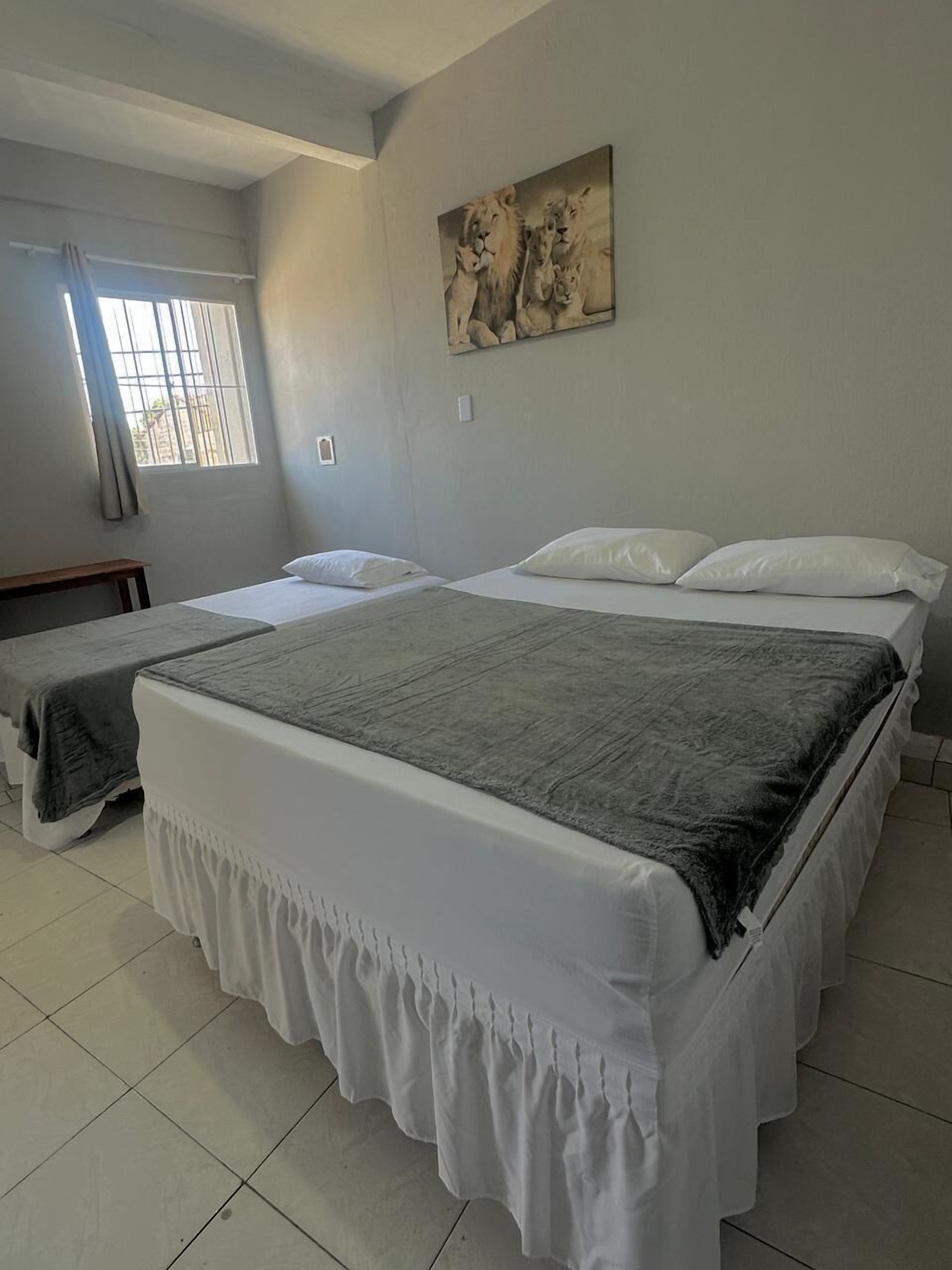 Une chambre d’hôtel avec deux lits, une fenêtre grillagée, un tableau au mur et un sol carrelé.