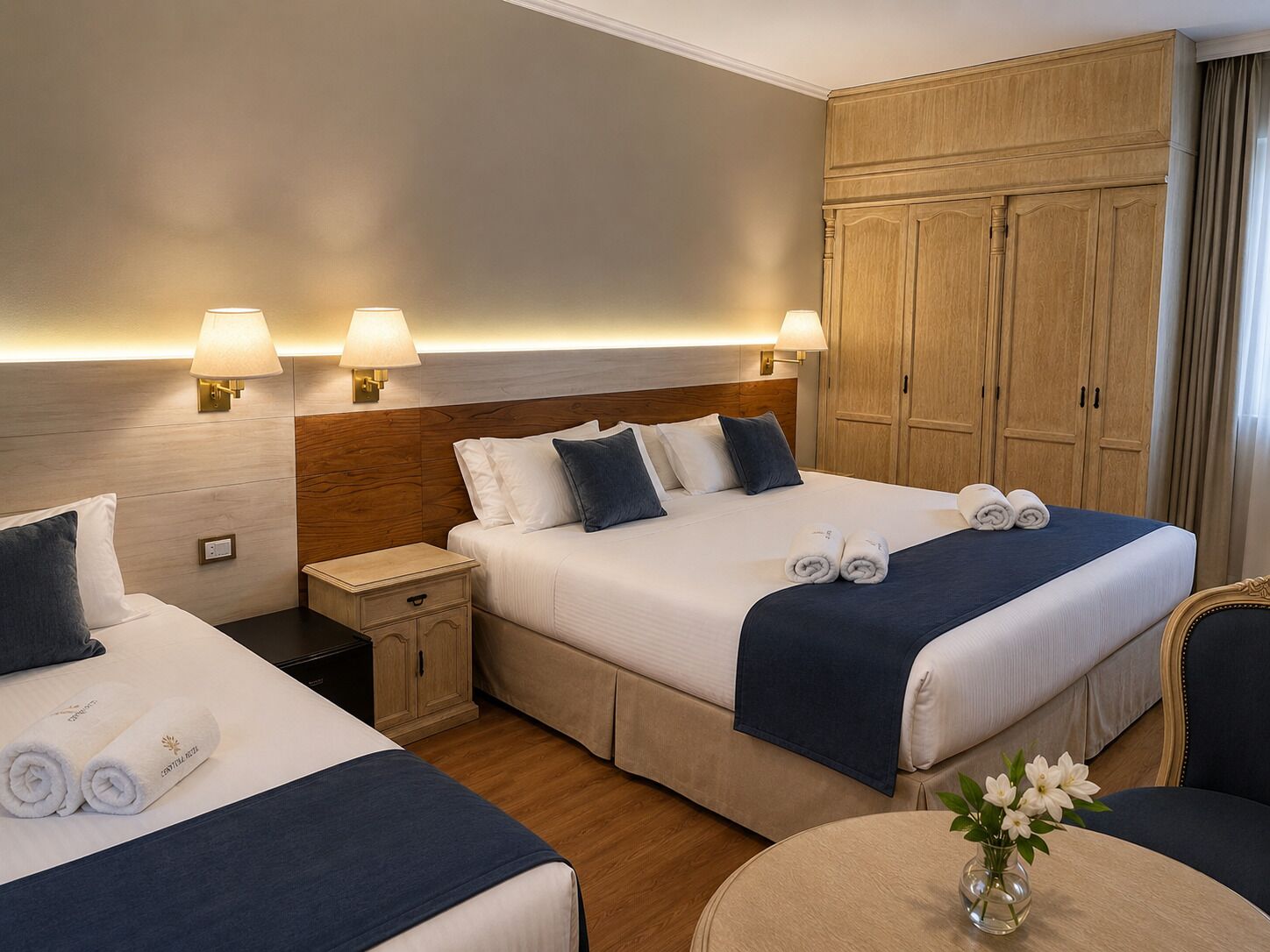 A hotel room with two beds, nightstands, a wooden wardrobe, and a small round table with a vase of flowers.