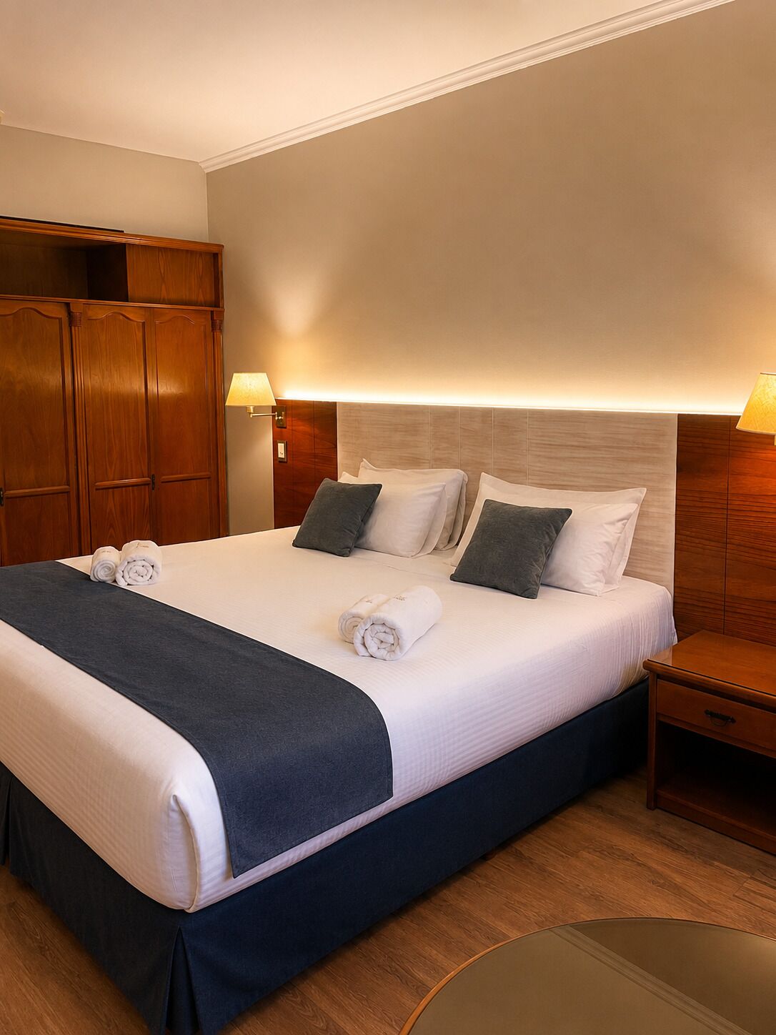A hotel room with a large bed, two bedside lamps, a wooden wardrobe, and a round table.