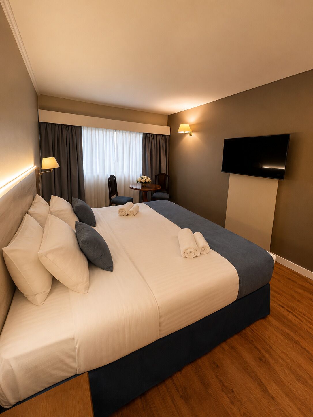 A hotel room with a large bed, a flat-screen TV mounted on the wall, and a small table with a chair.