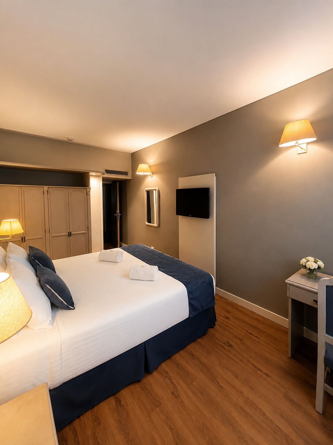 A hotel room with a bed, bedside lamps, a TV, a mirror, and a small table with a vase of flowers.
