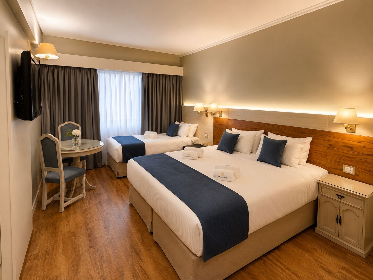 A hotel room with two beds, a small table with a chair, a TV, and a nightstand with a lamp.