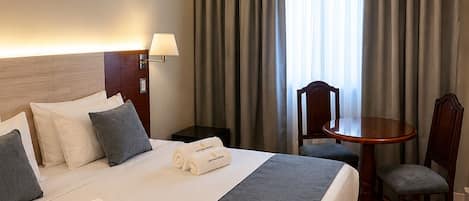 A hotel room with a large bed, a bedside table, a chair, a small table, and a lamp.