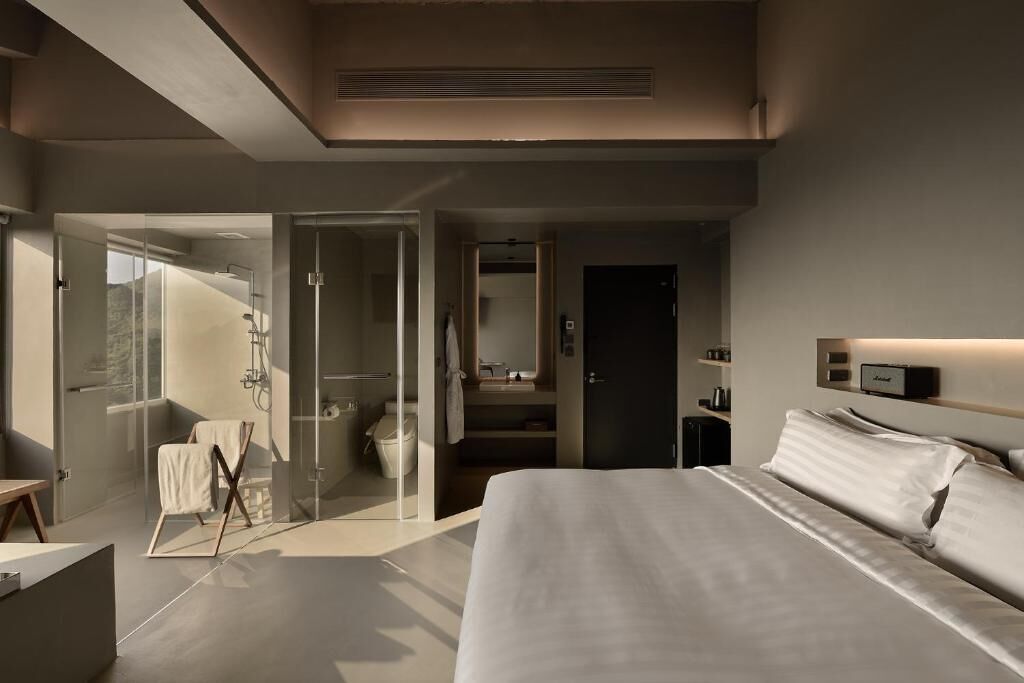 Ein modernes Hotelzimmer mit einem großen Bett, einer Duschkabine aus Glas, einem Schminktisch mit Spiegel und einem Nachttisch.