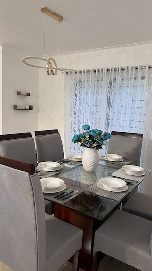 Un coin repas avec une table en verre, des chaises grises, un vase blanc orné de fleurs bleues et un luminaire moderne au plafond.