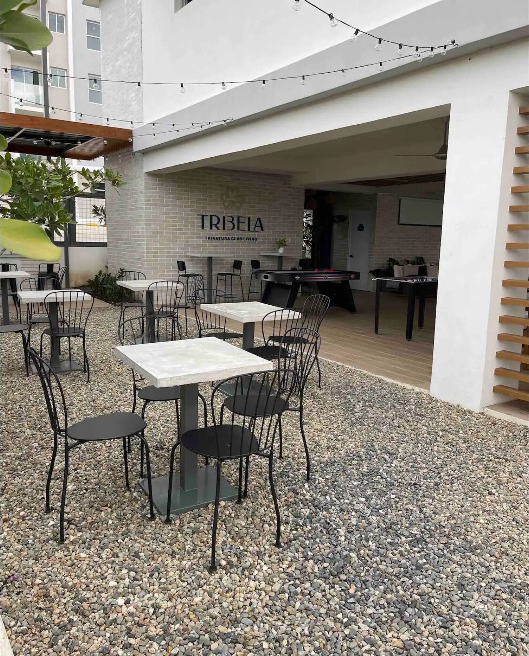 An outdoor seating area with tables and chairs, a pool table, and a sign for 'TRIBEIA TRINATURA CLUB LIVING.'