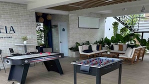 A pool table, a foosball table, and a sofa in a lounge area with a brick wall and plants.