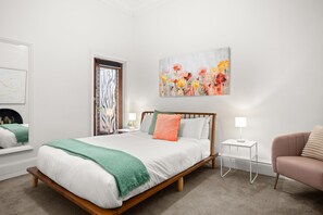 A bedroom with a bed, a nightstand, a chair, a painting, and a window with curtains. - Albertine House Pet Friendly Beachside Retreat (Albert Park)