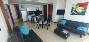 Ein Wohnzimmer mit schwarzem Sofa, Couchtisch und Fernseher. Es gibt eine Essecke mit Tisch und Stühlen.