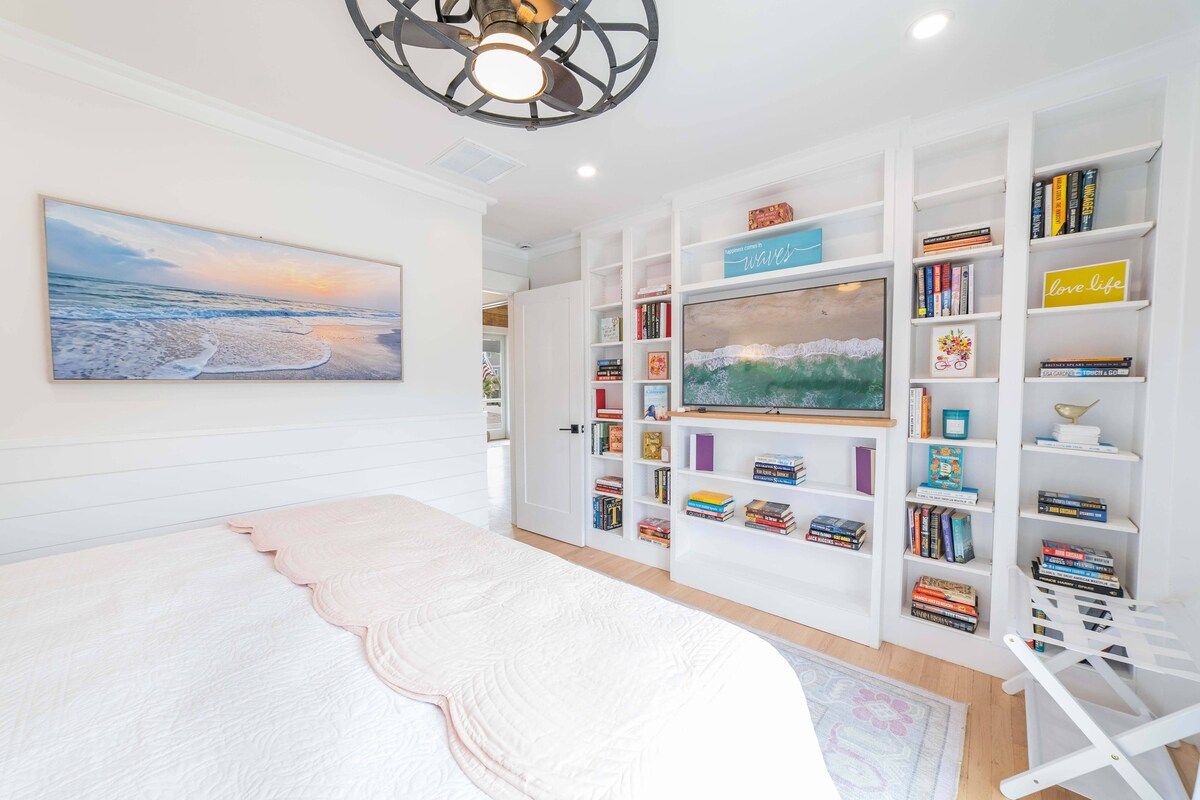 A bedroom with a large bed, a bookshelf, a television, and a painting of a beach scene.