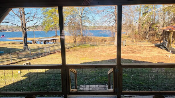 Une vue à travers la fenêtre d’un bâtiment doté d’une terrasse en bois, d’une zone délimitée par une clôture et, au loin, d’une étendue d’eau.