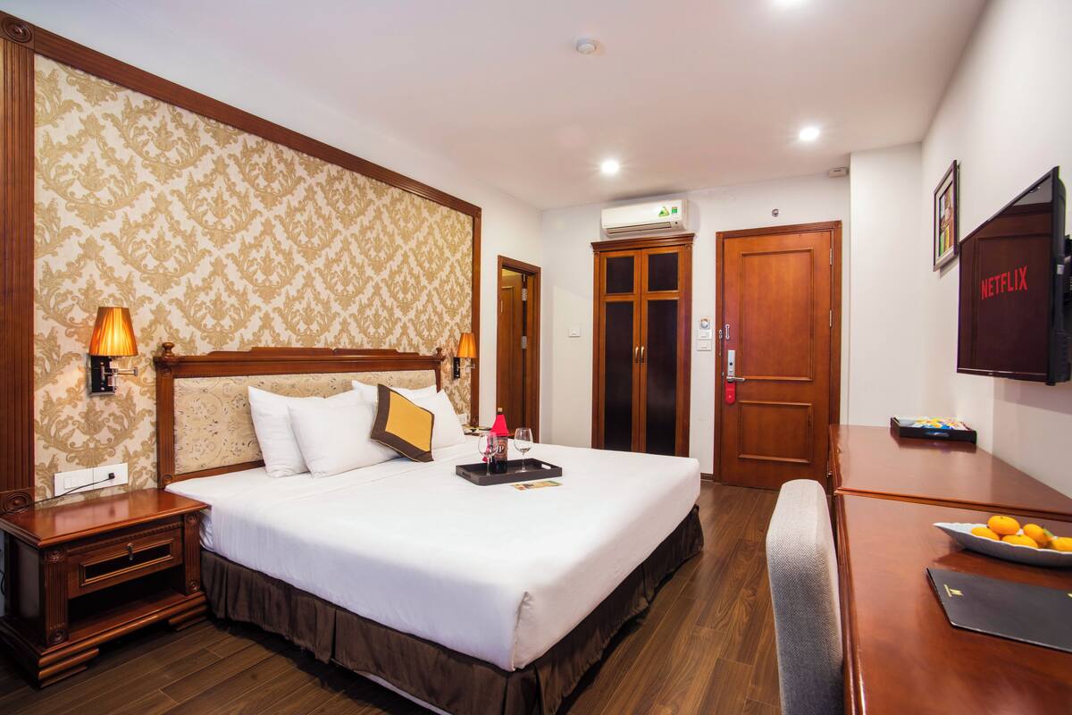A hotel room with a large bed, wooden headboard, and a bedside table with a lamp. There is a desk with a laptop and a bowl of fruit.
