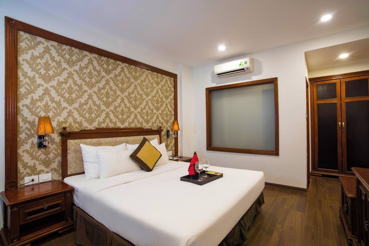 A hotel room with a large bed, a nightstand, a lamp, an air conditioning unit, and a wardrobe.