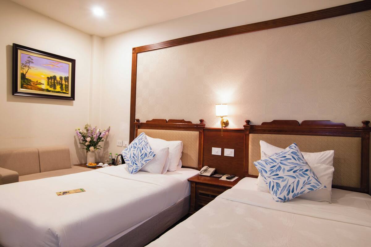 A hotel room with two beds, a nightstand, a lamp, a vase with flowers, and a framed picture on the wall.