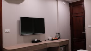 Habitación de hotel con televisor de pantalla plana montado en la pared, un escritorio de madera con cafetera y una puerta de madera cerrada.