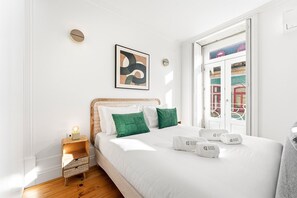 A hotel room with a bed, a bedside table, a wall clock, a framed abstract painting, and a window with a view of buildings. - GuestReady - Charming studio in Porto's heart (Porto)