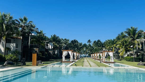 Une piscine d’un complexe hôtelier, avec une eau d’un bleu limpide, entourée de palmiers et de bâtiments modernes.