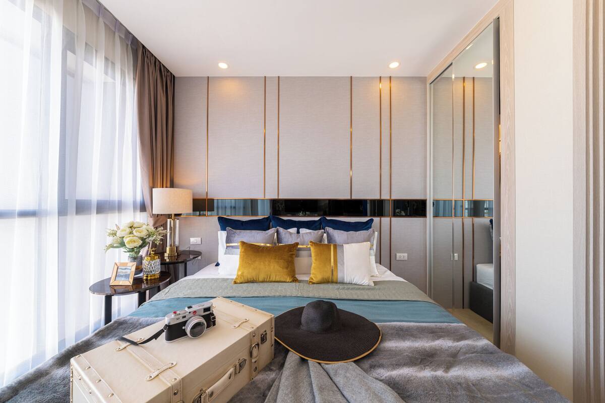 Una habitación de hotel con cama, mesita de noche, lámpara, maleta y sombrero.