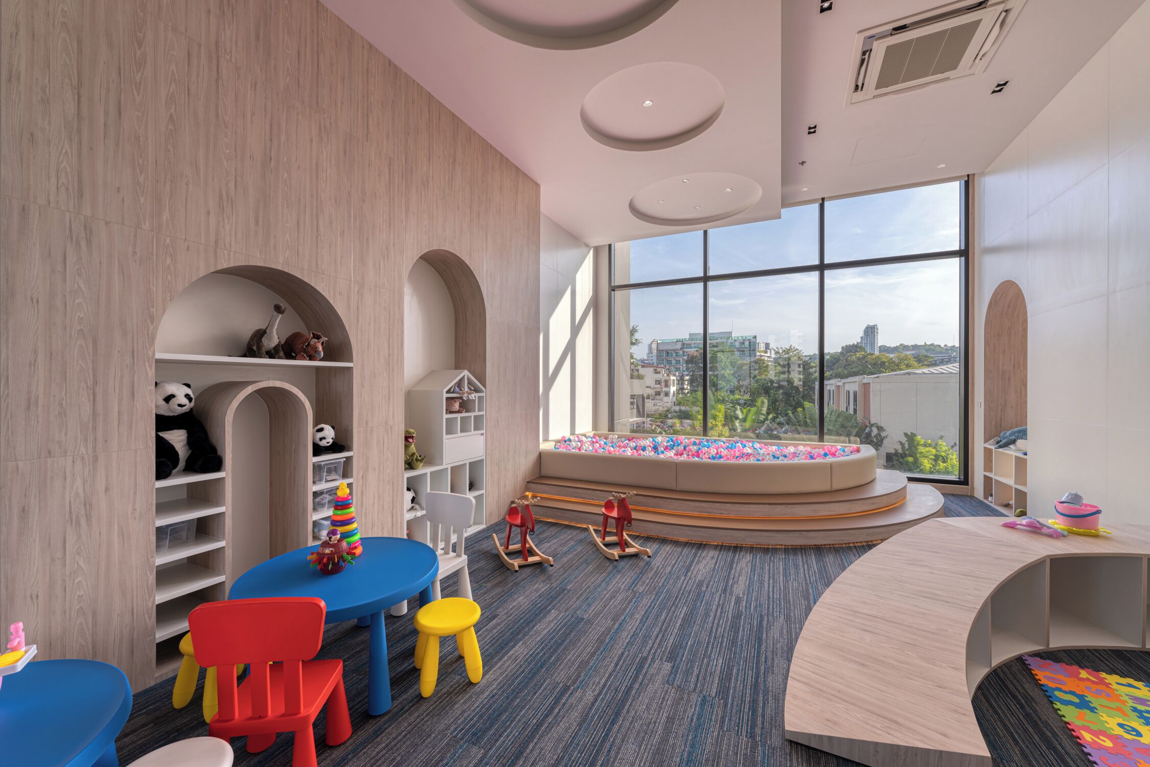 A children's playroom with a large window, wooden shelving, and colorful play furniture.