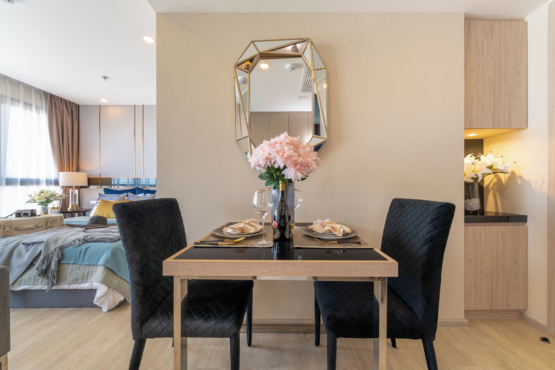 A dining area with a table set for two, a mirror above, and a bed with a view in the background.