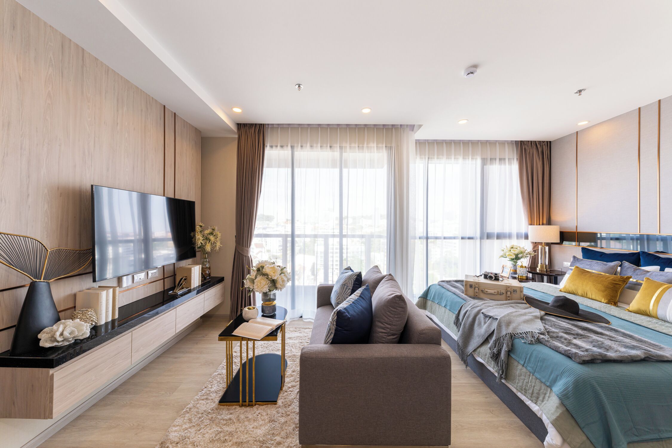 A modern hotel room with a flat-screen TV, a sofa, a bed with multiple pillows, a bedside table, and a large window.