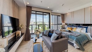 A modern hotel room with a large flat-screen TV, a sofa, a bed with multiple pillows, and a view of the cityscape through large windows. - The Panora Pattaya by PH (Pattaya)