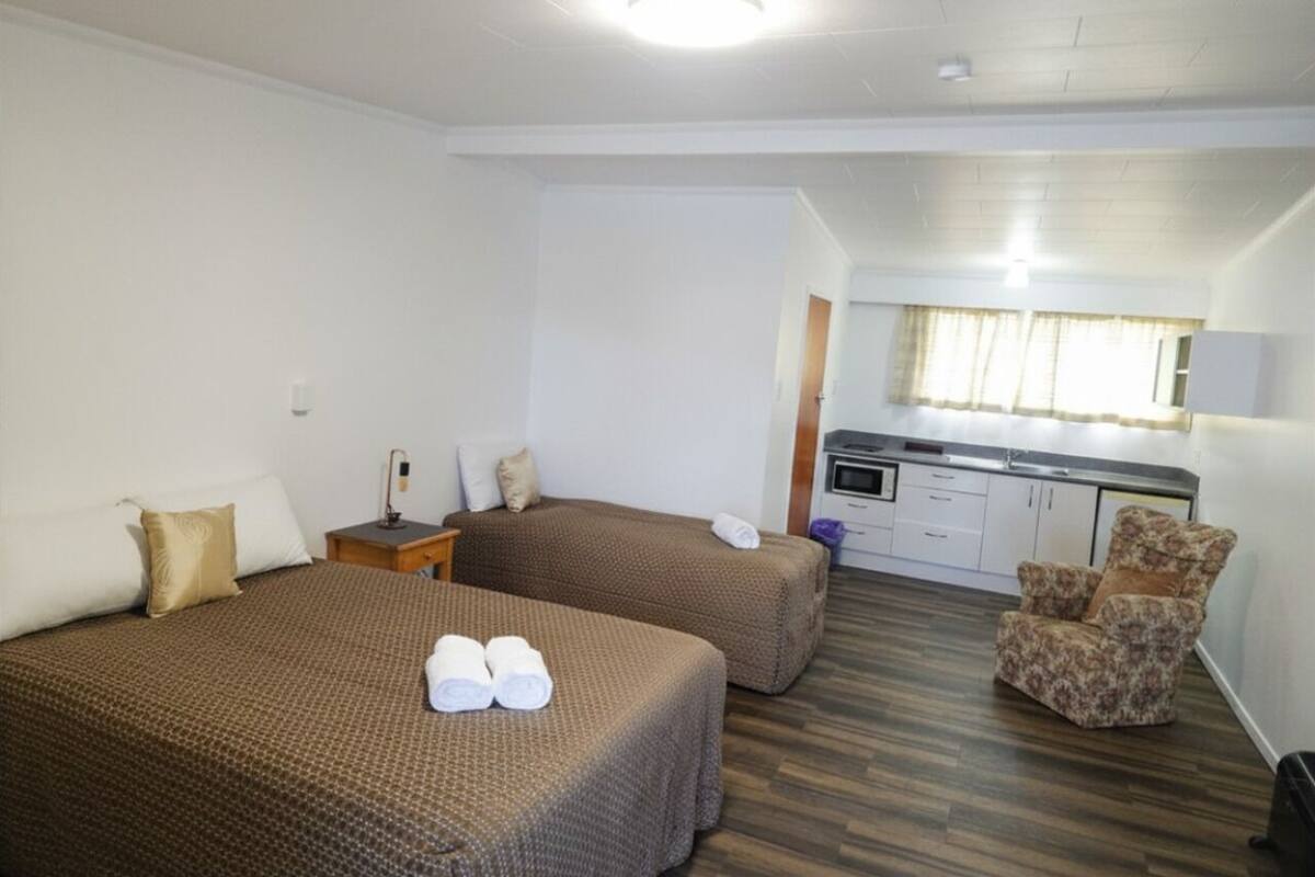 A room with two beds, a chair, a kitchenette, and a window.