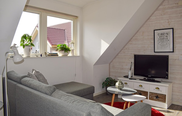 A modern living room with a grey sofa, a TV, a small table, and a window with plants.